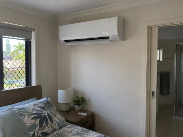 Wall-mounted split system above a bedroom bed, with window, bedside table and open louvre in Broadbeach